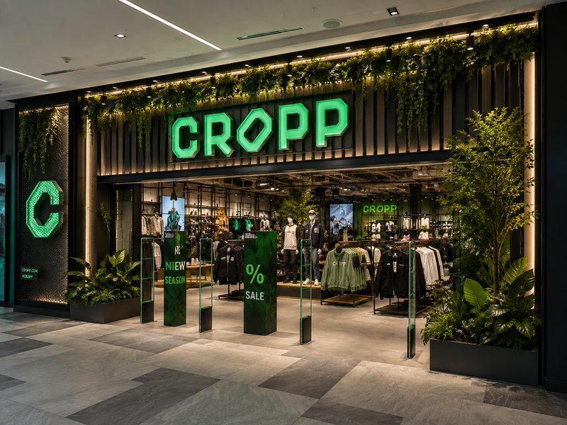 Yeşil bitkilerle çevrili, aydınlatmalı Cropp mağaza girişi.